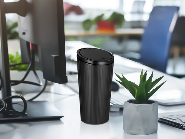 Black cylindrical travel mug or thermos on desk beside computer monitor and small potted plant in office setting.