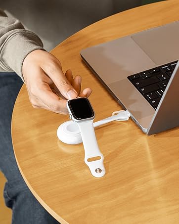 Smartwatch being placed on white charging dock next to laptop on wooden surface. Hand visible positioning the watch on charger.
