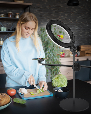 Woman in blue sweater chopping vegetables on cutting board in kitchen. Large circular light fixture illuminates workspace. Vegetables and cooking utensils visible on counter.