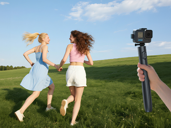 Handheld camera gimbal with action camera mounted, capturing two women running in a grassy field. Blue sky with clouds in background.