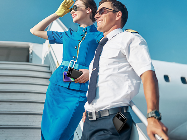 Two people in airline uniforms standing near an aircraft. Woman in blue dress, man in white shirt. Both wearing sunglasses and looking off-camera.