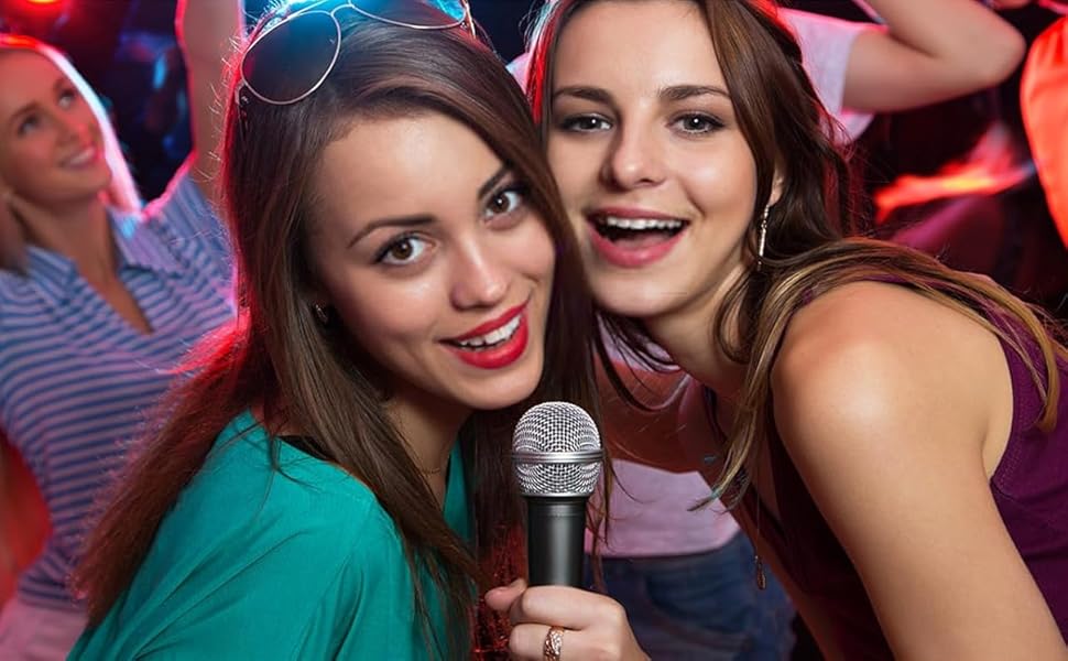 Handheld microphone in a colorful party or karaoke setting with red lighting and crowd in background.