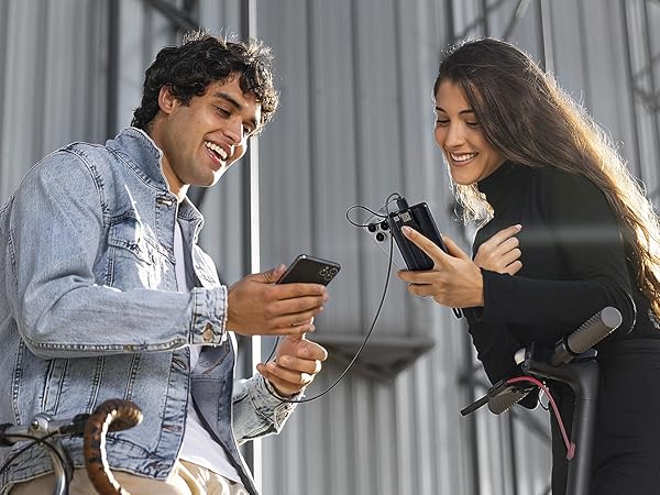 Small handheld microphone attached to smartphone. Woman holds microphone while man holds phone, both smiling. Outdoor setting with metal background.
