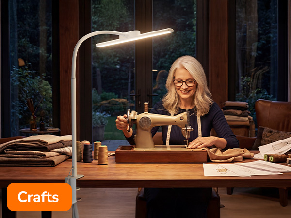 Text reads 'Crafts.' Scene shows person using sewing machine at wooden desk illuminated by modern LED desk lamp against dark window background.