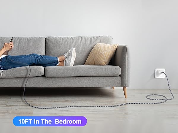 Gray sofa with person lounging, using smartphone connected to long charging cable extending to wall outlet. Text overlay: '10FT In The Bedroom'.
