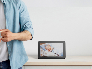 Rectangular digital display device on a table, showing an image of a baby. Person's arm visible in blue shirt.