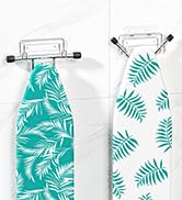 Two white hangers displaying ironing board covers with tropical leaf patterns. One has teal leaves on white, the other white leaves on teal background.