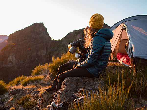 Person in blue jacket and yellow hat sitting on rock, holding mug near orange tent. Mountainous landscape at sunset or sunrise.