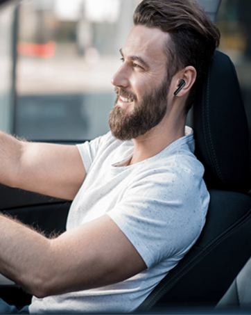 Man with beard wearing white t-shirt and wireless earbuds, seated in vehicle, looking to the side with a smile.