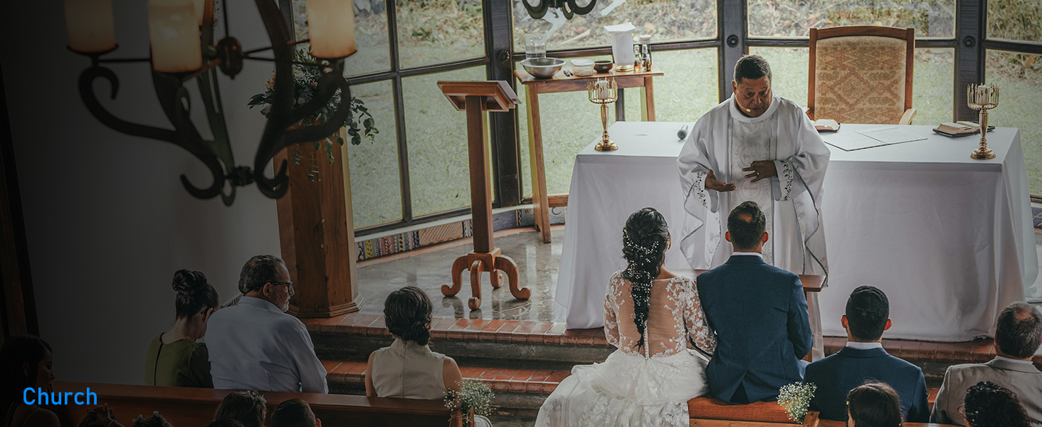 A religious ceremony taking place in a chapel setting with wooden beams and natural lighting, with attendees seated in rows.