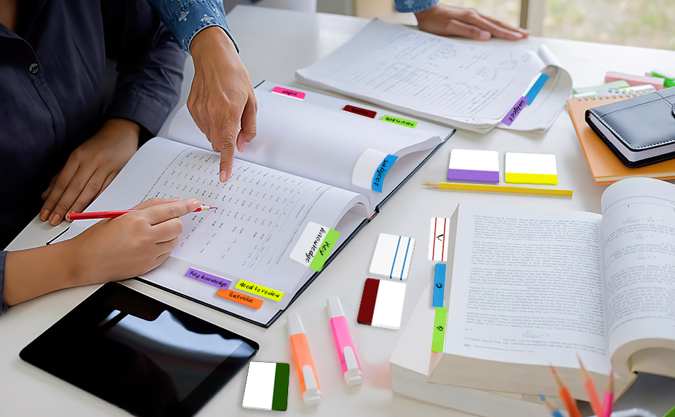 Desk with open notebooks, colorful sticky notes, and highlighter pens. Hands visible writing or studying, suggesting academic or office setting.