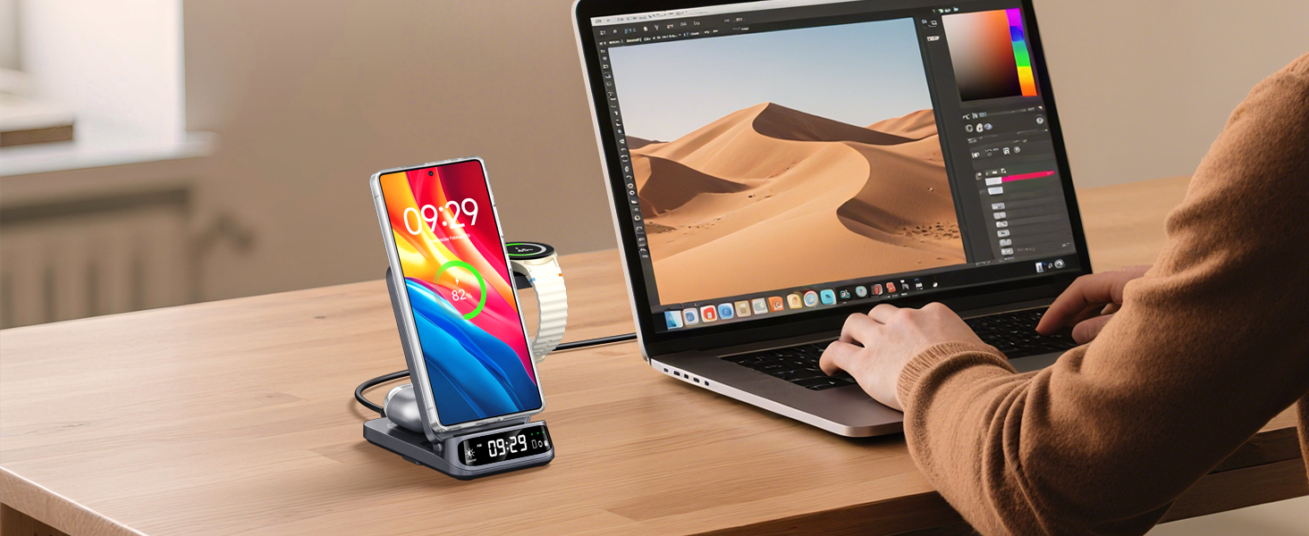 Person using laptop with desert image on screen. Smartphone in charging dock displays colorful wallpaper. Both devices on wooden desk, showcasing mobile workstation setup.