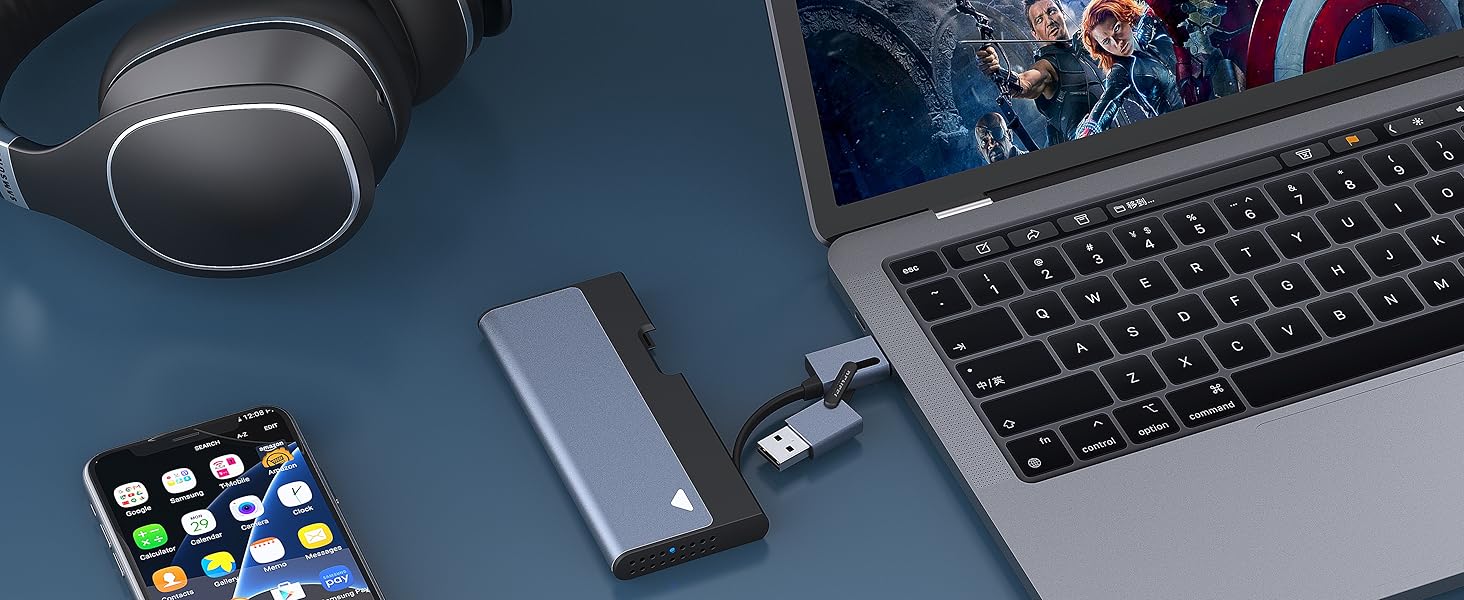 Tech accessories on dark surface: laptop, smartphone, wireless earbuds in case, and portable storage device or adapter connected to laptop.