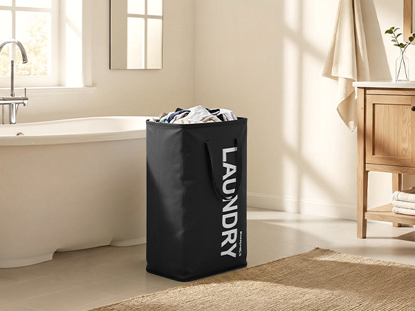 Text reads 'LAUNDRY'. Black cylindrical laundry hamper with white text positioned in a sunlit bathroom corner near a freestanding bathtub and wooden furniture.