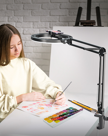 Adjustable desk lamp with magnifying glass lens, used by person painting. Features flexible arm and clamp for desk attachment.