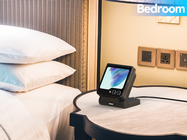 Small touchscreen device on charging dock, placed on bedside table. Wicker headboard visible with white pillows. Wall outlets and 'Bedroom' text in background.