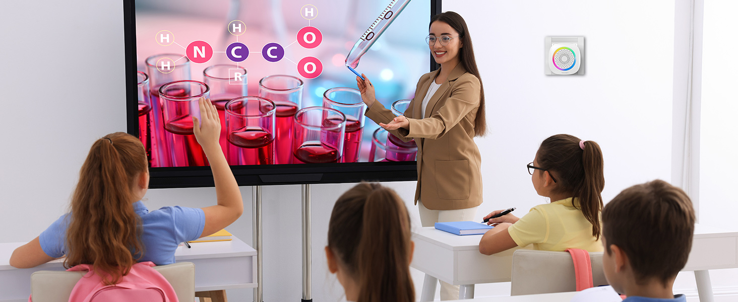 Classroom setting with students and teacher using an interactive display. The screen shows colorful graphics related to a lesson, while the teacher gestures towards it.