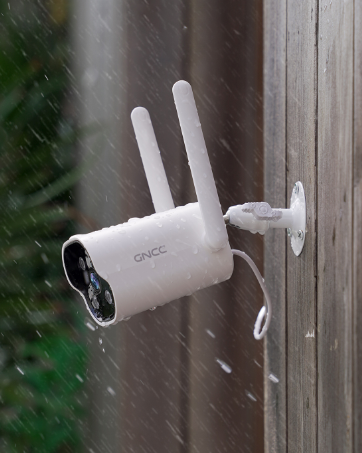 White outdoor security camera with two antennas mounted on a wooden surface. Camera has visible infrared sensors and is exposed to rain or snow.