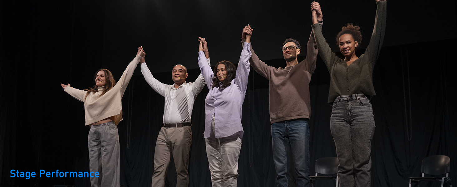 Stage performance scene showing performers in casual attire with raised joined hands against dark background.