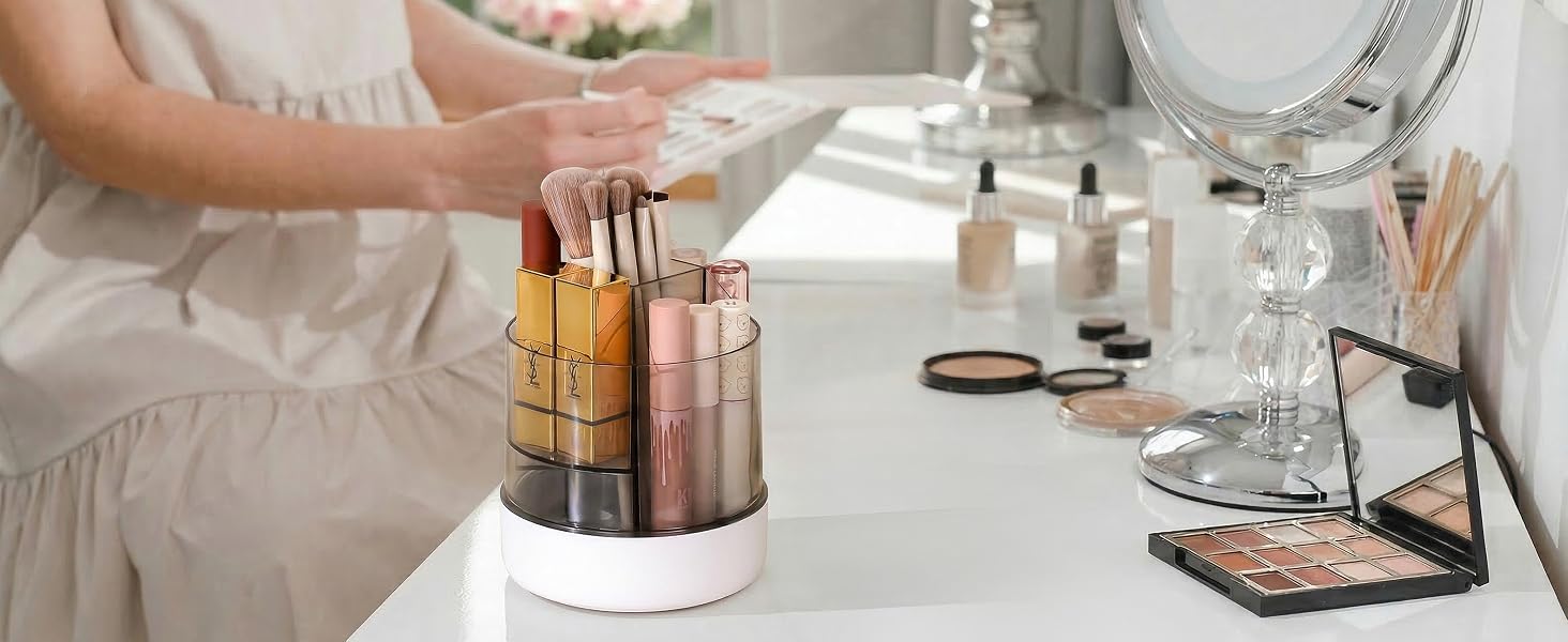 Collection of cosmetic products displayed on white surface. Multiple metallic tubes and containers arranged in cylindrical holders. Glass bottles and makeup palettes visible in minimalist, elegant setup.