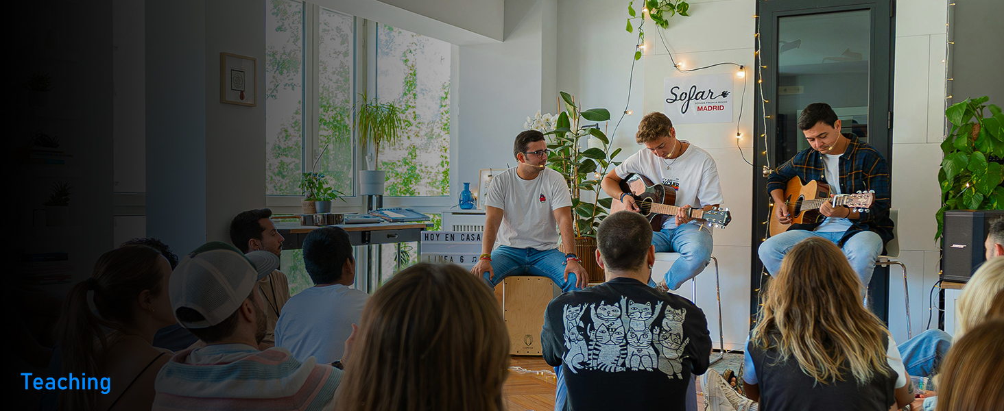 A group performs acoustic music in an indoor setting with plants and white columns, audience seated and watching.