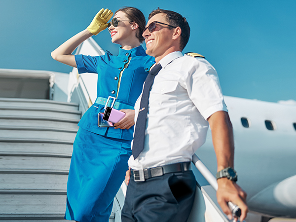Two airline crew members on aircraft stairs. Woman in blue uniform shielding eyes, man in white shirt holding luggage.