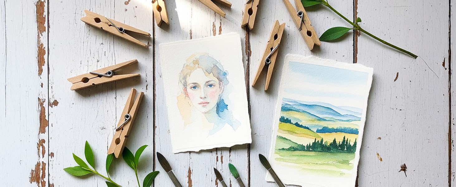 Gallery wall display showing watercolor artwork and photographs arranged on distressed white wooden wall with green leaf accents.