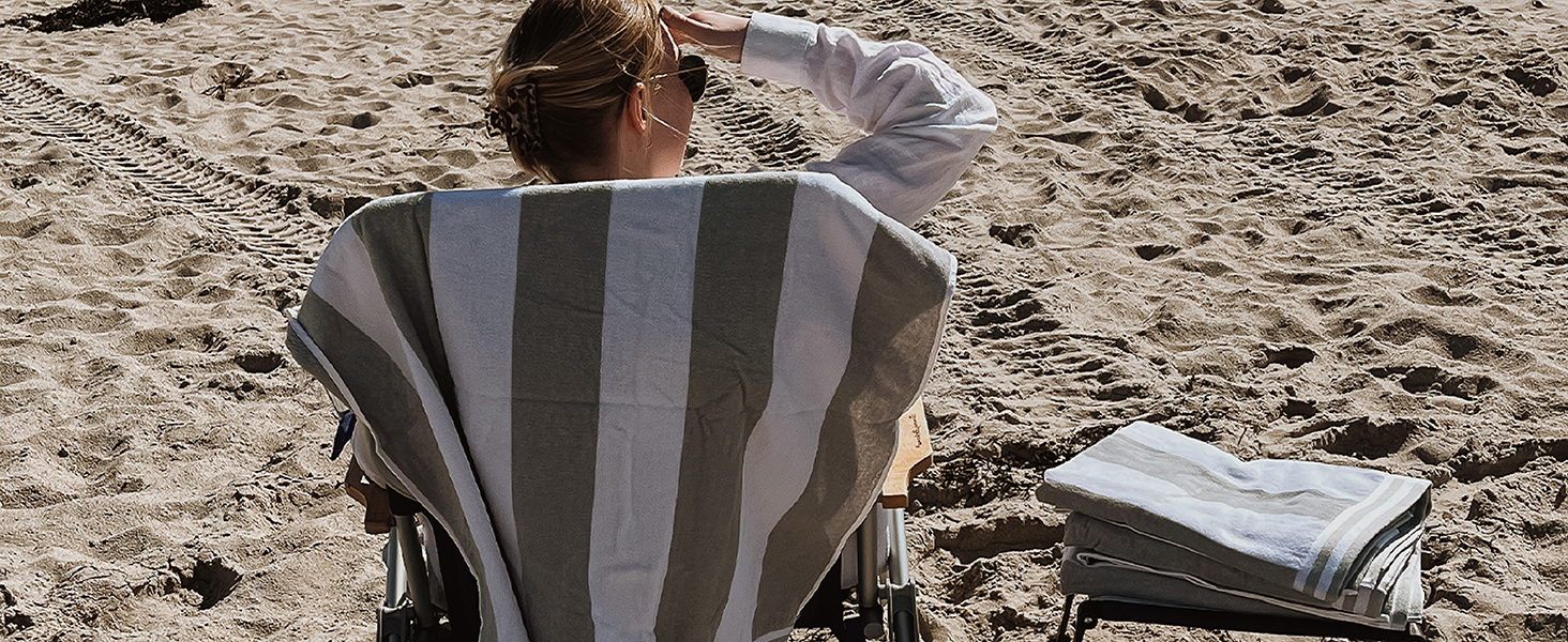 A woman sitting down at the beach with her beach towel hanging from the back of the chair.