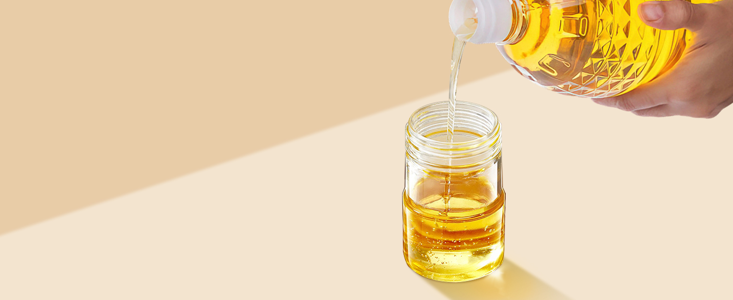 Hands pouring golden liquid, likely cooking oil, from a large plastic bottle into a small glass jar on a beige surface.