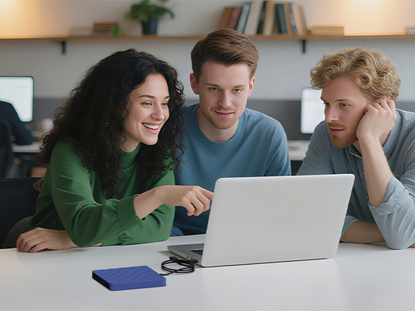 Multiple people gathered around a laptop in a casual indoor setting, looking engaged with the screen content. Room has bookshelves and natural lighting in background.