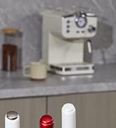 Compact espresso machine with white finish on marble countertop. Coffee cup and storage container visible. Three bottle tops in foreground.