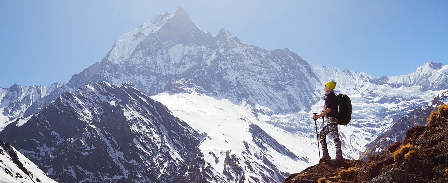 Hiker with backpack standing on rocky terrain, facing snow-capped mountain peaks against clear blue sky.