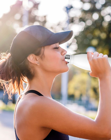 Athletic woman in dark activewear and cap drinking from a water bottle outdoors. Sunlit background with trees suggests park or nature setting.