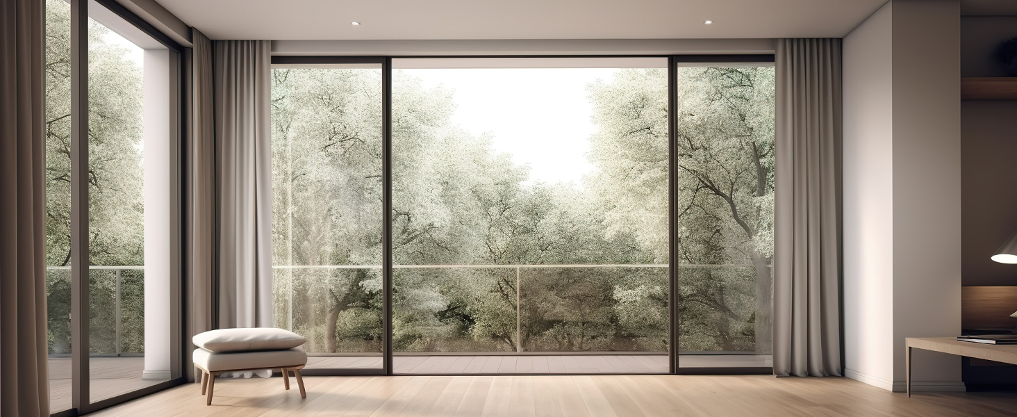 Modern interior with large floor-to-ceiling windows overlooking green foliage. Minimalist furniture including chair visible. Natural light illuminates the room.