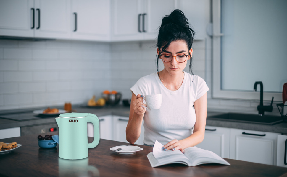 Mint green electric kettle on kitchen counter. Woman in white shirt reading book and holding mug in background, demonstrating product use in home setting.