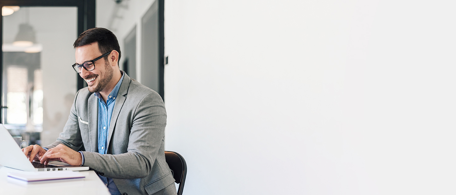 Man in business casual attire smiling while working on a laptop at a desk in an office setting.