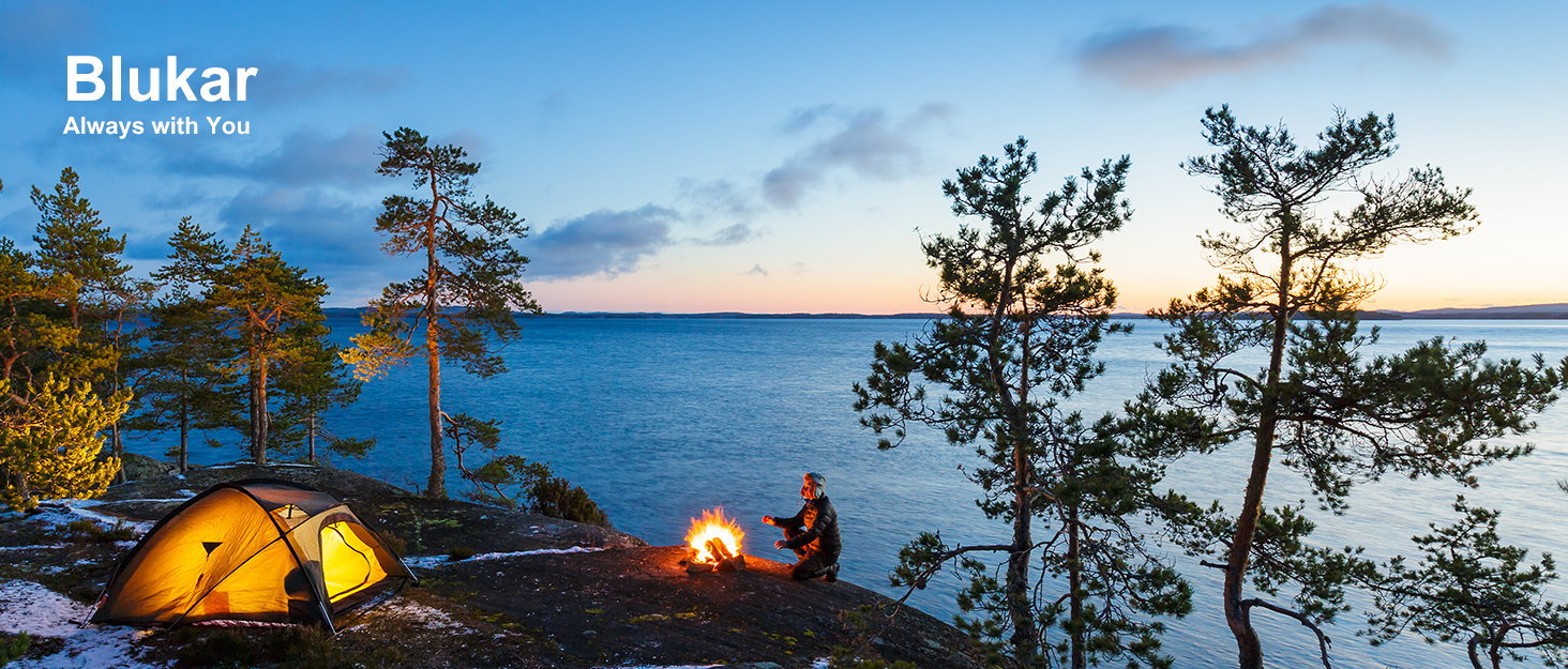 Camping scene with yellow tent and campfire on rocky shore. Pine trees frame view of calm lake at sunset.