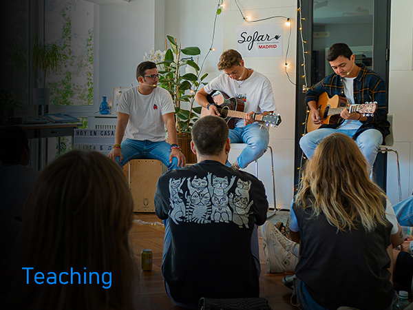 Indoor music performance setting with acoustic guitars and performers in casual attire, audience seated in foreground.