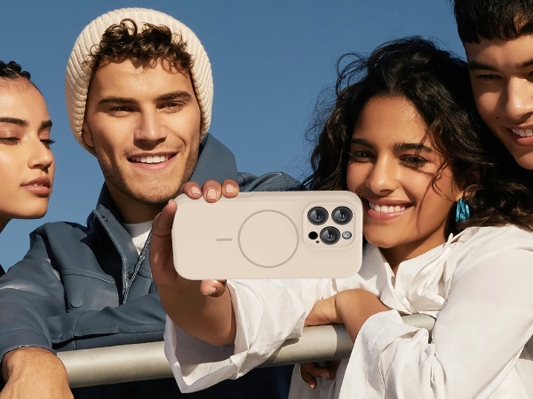 Four diverse young people outdoors against blue sky, holding white smartphone displaying camera interface. Group posed casually while showcasing mobile device.