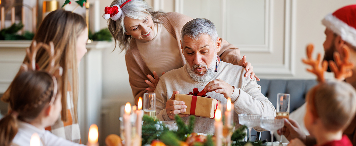 Family gathering around a Christmas table. Elderly man receiving a gift, surrounded by others in festive attire.
