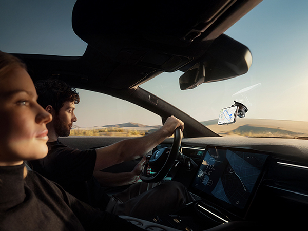 Interior view of a modern car dashboard with large touchscreen display. Steering wheel and digital instrument cluster visible. Sunset landscape through windshield and sunroof.