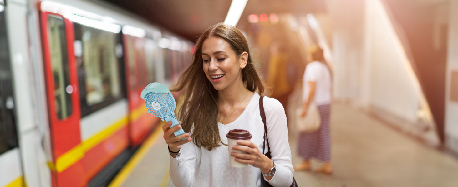 Woman holding a turquoise handheld fan and smartphone on a subway platform with a train visible in the background.