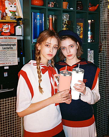 Two young women in casual outfits holding beverages in a dimly lit, retro-styled interior with shelves and decorative items visible in the background.