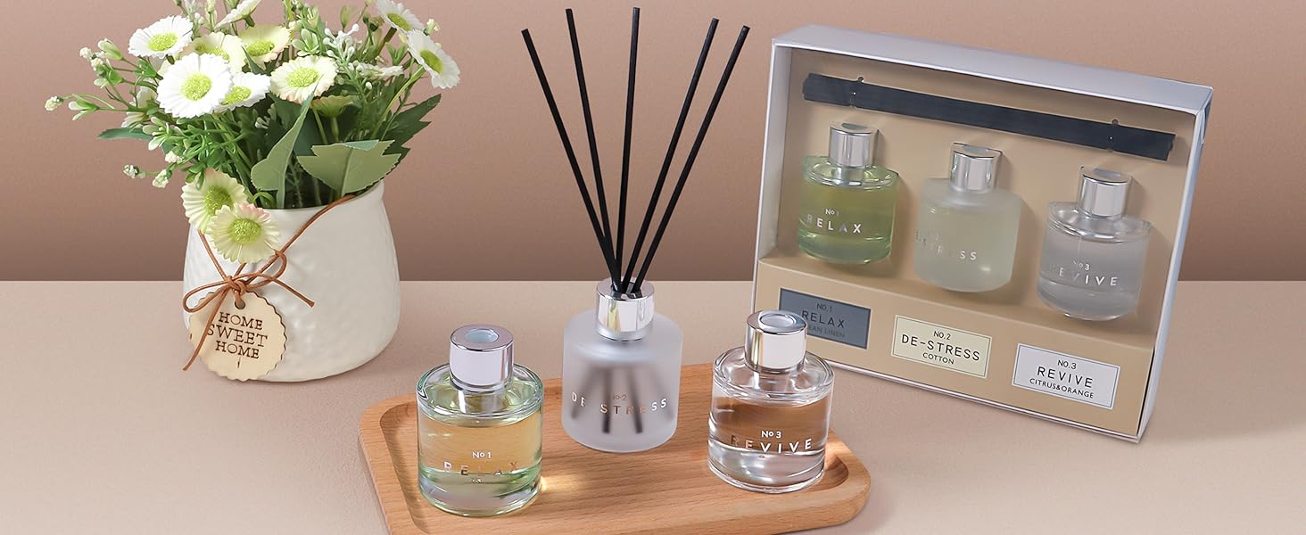 Home fragrance display on wooden tray featuring reed diffuser, scented candles, and decorative packaging, alongside small potted white flowers in burlap-wrapped container.