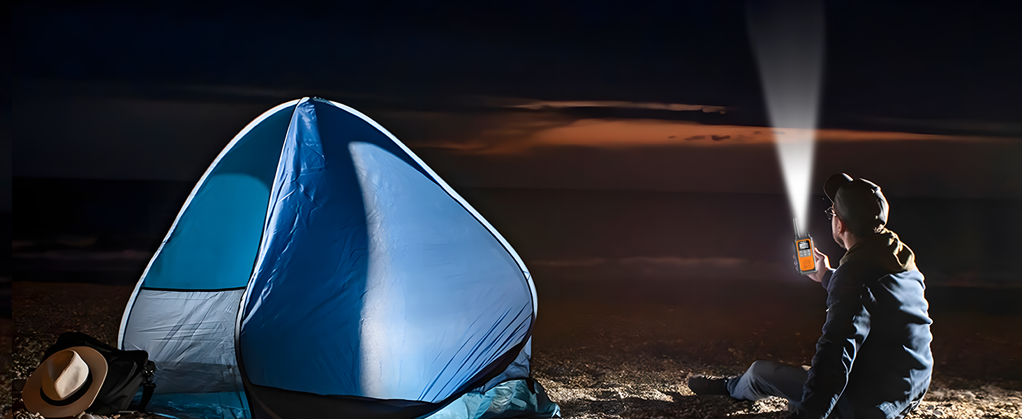A camper sits by tent at night using walkie talkies with its flashlight feature to light darkness