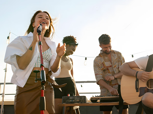 Outdoor music performance scene with performers using microphone, turntable, and acoustic guitar. String lights visible in background, suggesting casual or beachside setting.