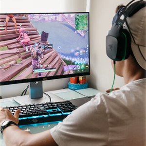 Person wearing gaming headset seated at desk with computer monitor, keyboard, and controller visible. Screen displays colorful game interface.