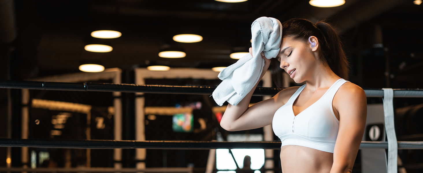 Athletic woman in white sports bra wiping sweat with a white towel in a gym setting with overhead lighting.