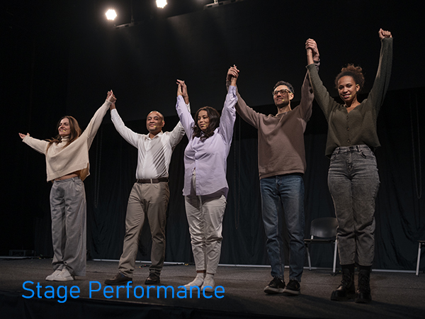 Performers on a dark stage taking a bow with raised joined hands at curtain call, wearing casual attire against black backdrop.