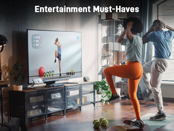Large TV displaying fitness content as two people exercise in a living room. Entertainment setup includes gaming accessories and plants on a media console.